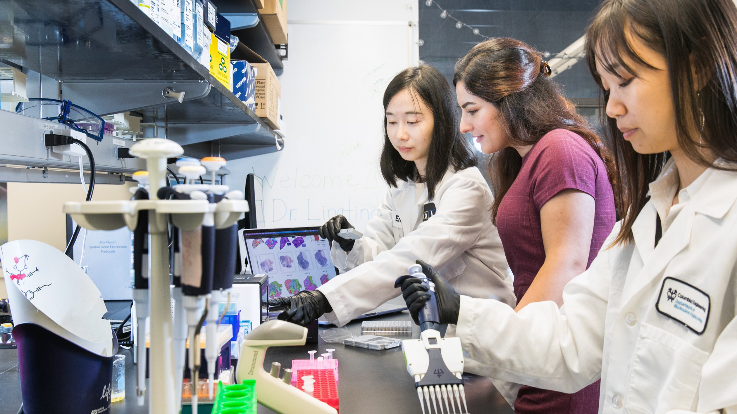 Three researchers in lab coats and gloves work at a laboratory bench with pipettes and samples while another person stands beside them, looking on.