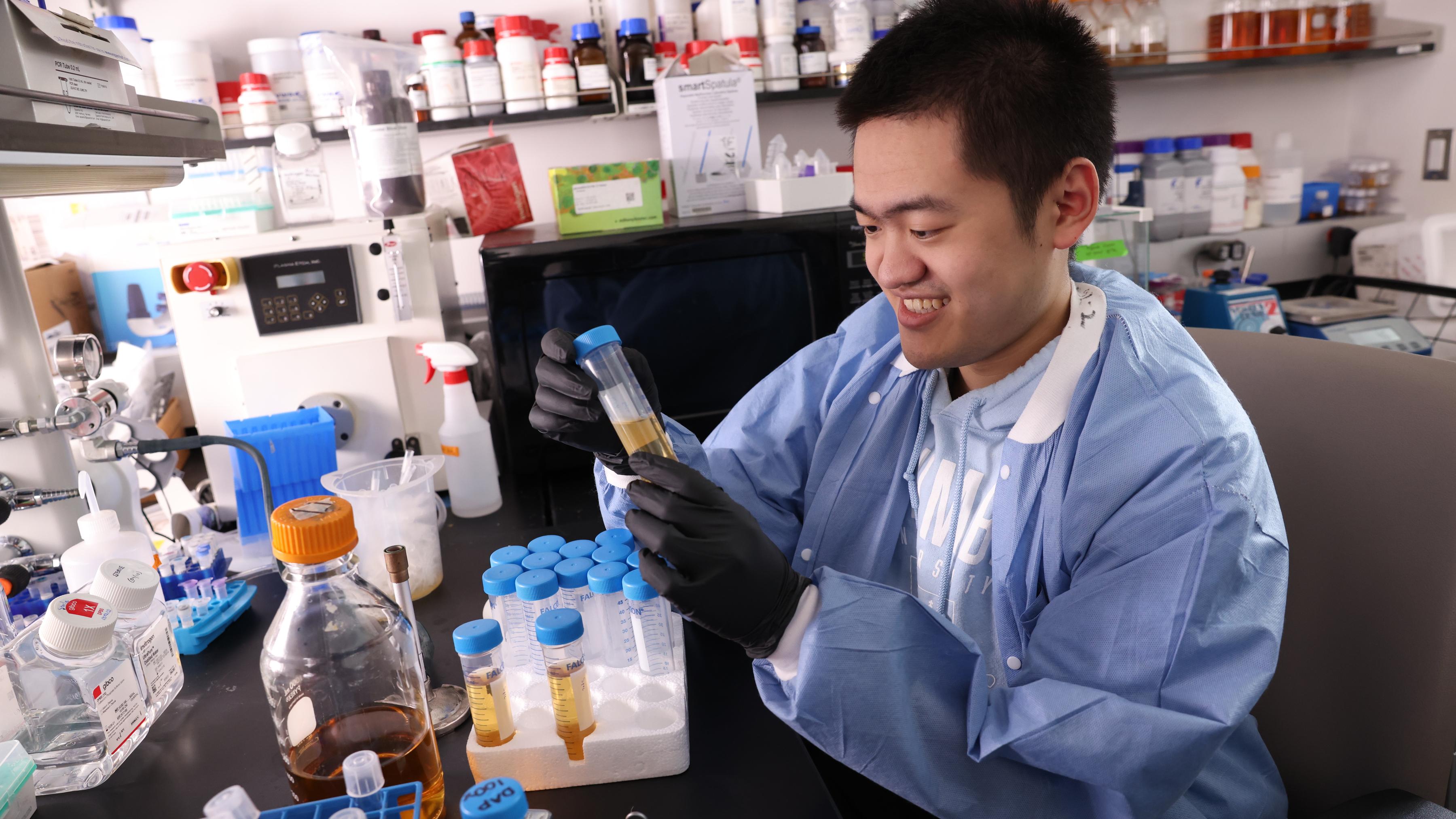 A researcher wearing gloves and a blue protective gown smiles while examining a sample tube at a lab bench filled with bottles, test tubes, and equipment.