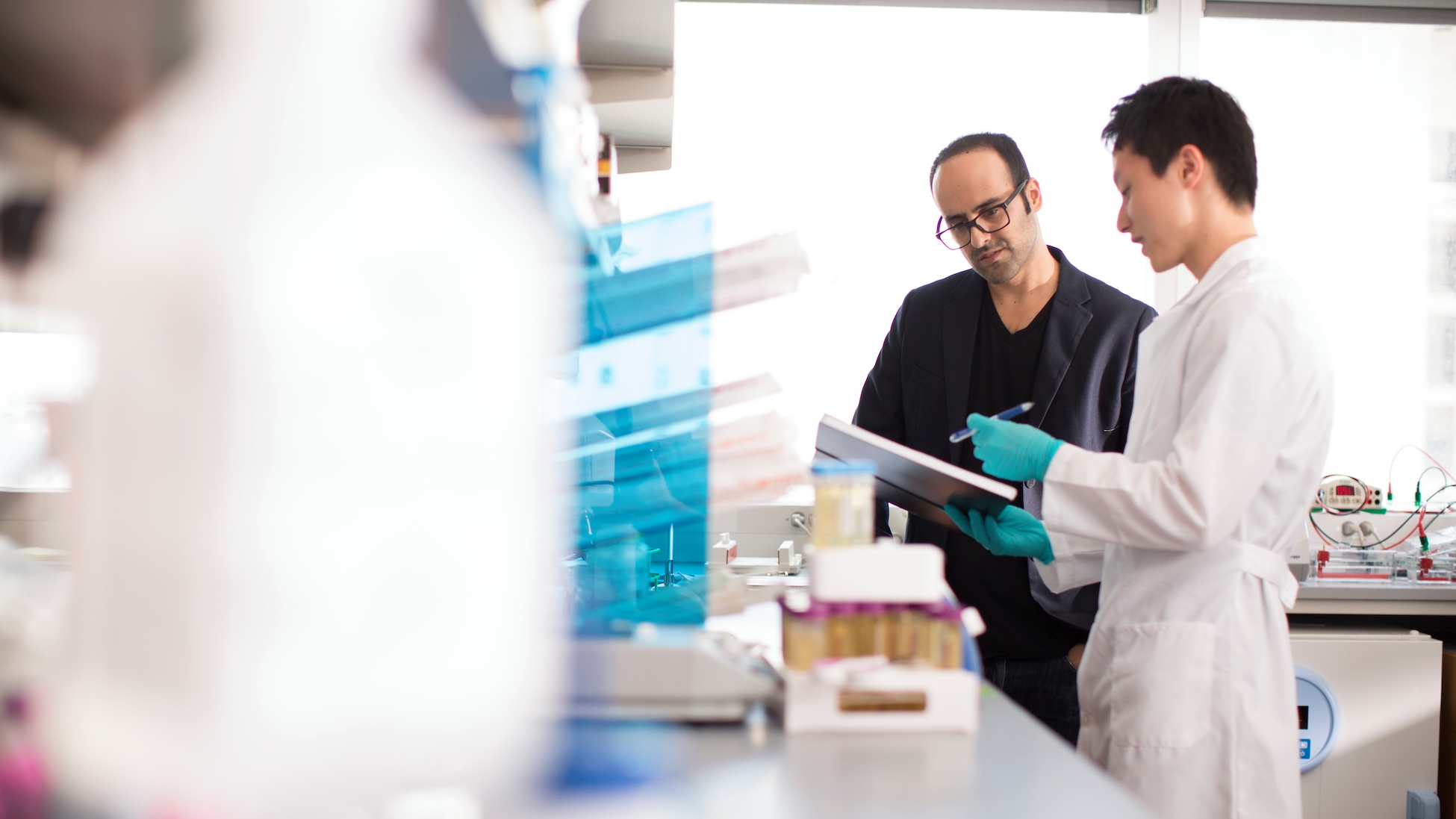 In a bright laboratory, a student in a white lab coat and blue gloves points to notes on a clipboard while discussing them with a professor; lab equipment and sample containers sit on the bench in the foreground.