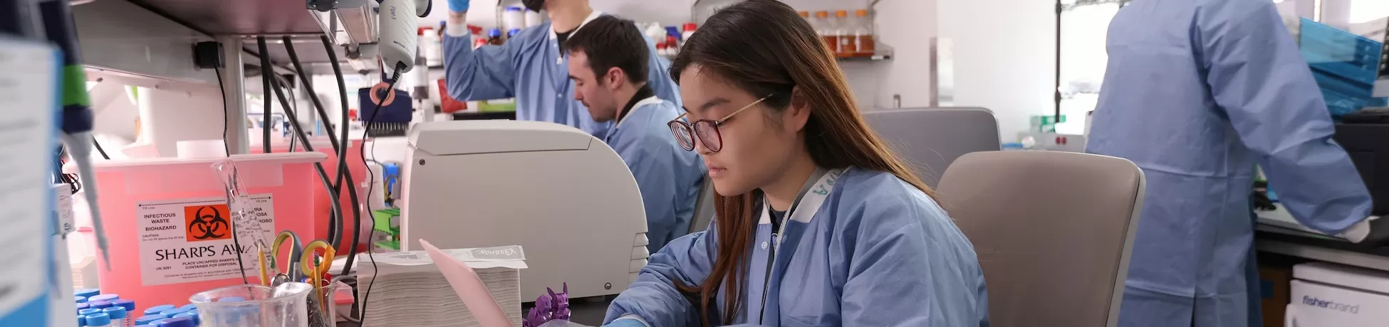 Research lab with four Columbia Engineering graduate students in blue lab coats and gloves; one sits at a bench working on a laptop while others handle equipment and samples near shelves of bottles and a sharps container.
