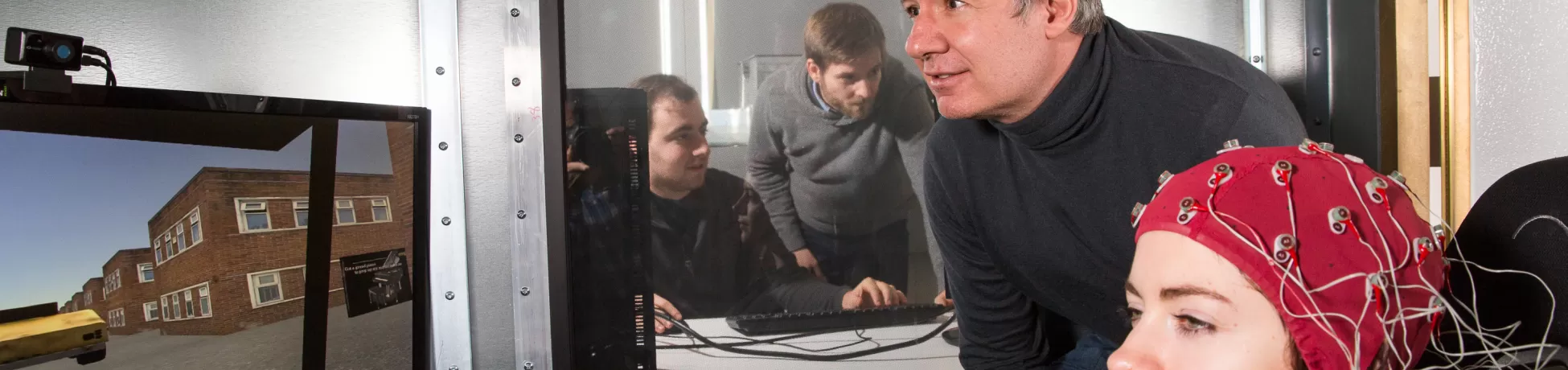 A participant wearing an EEG cap sits in a lab while a researcher watches a monitor displaying a virtual environment, with additional team members observing through a window.
