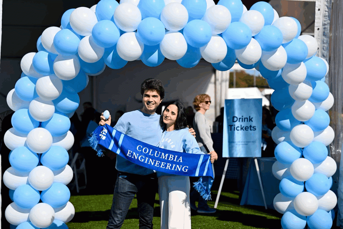 Two people smiling for a photo and holding a scarf that reads "Columbia Engineering" underneath a blue and white balloon arch 
